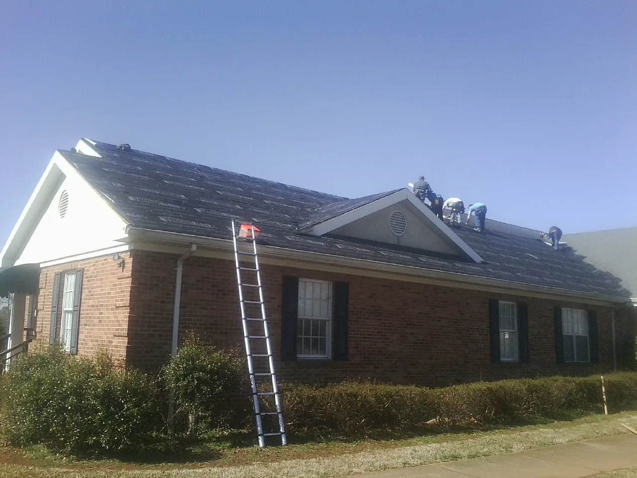 Crew tearing shingles off a brick ranch roof with a ladder set — Metal Roof Repair work by our crew in Lindsay
