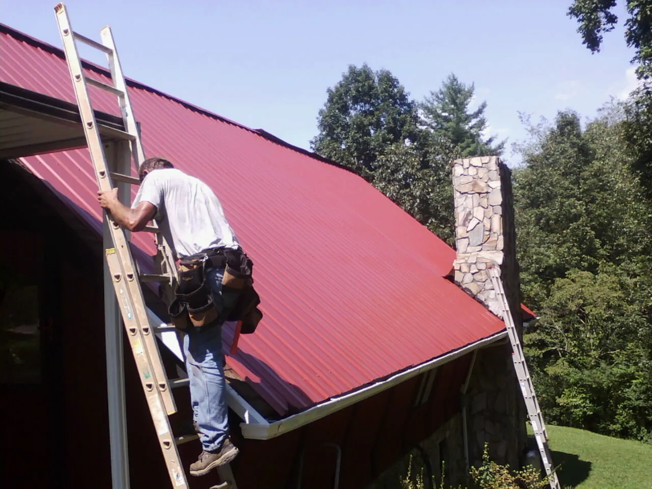 Roofer on a ladder beside a red metal roof and stone chimney — Soffit & Fascia Repair by Roofing Brothers in Lindsay