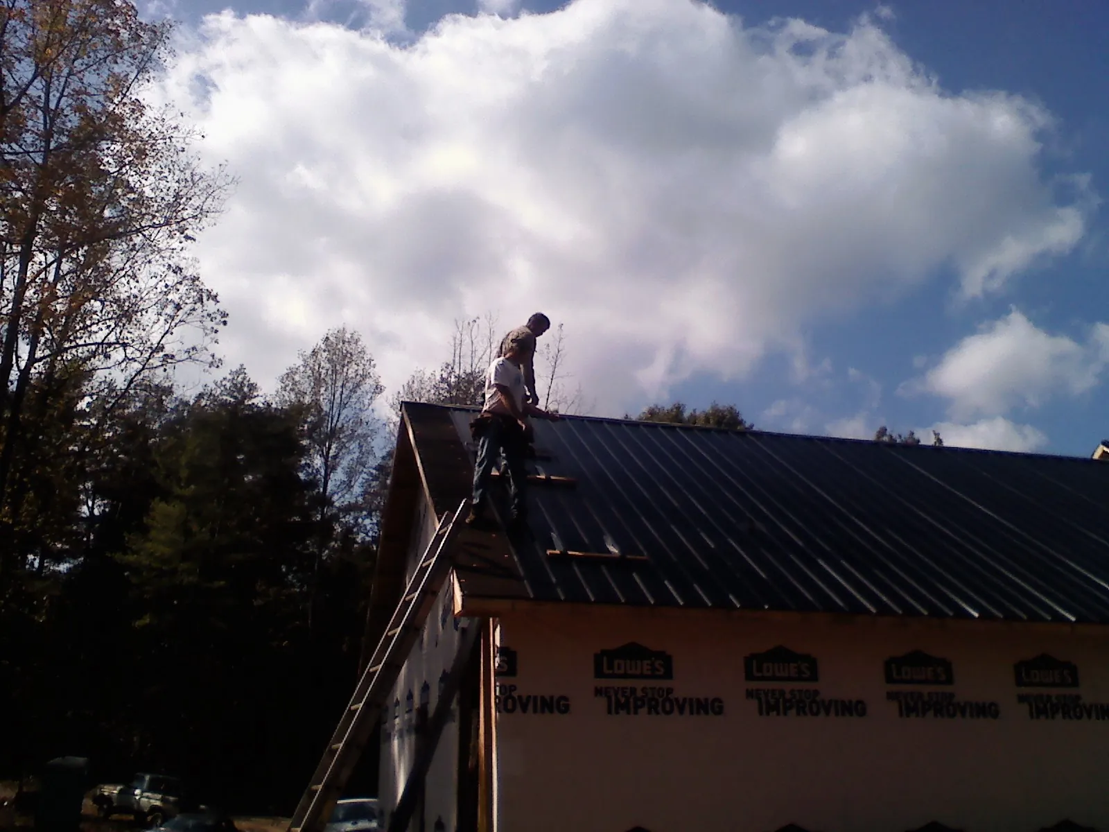 Roofer fastening dark metal panels high against a cloudy sky, from a Roof Inspection job in Lindsay
